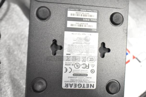Netgear device bottom view showing model GS105NA and serial number. Features rubber feet and mounting slots.