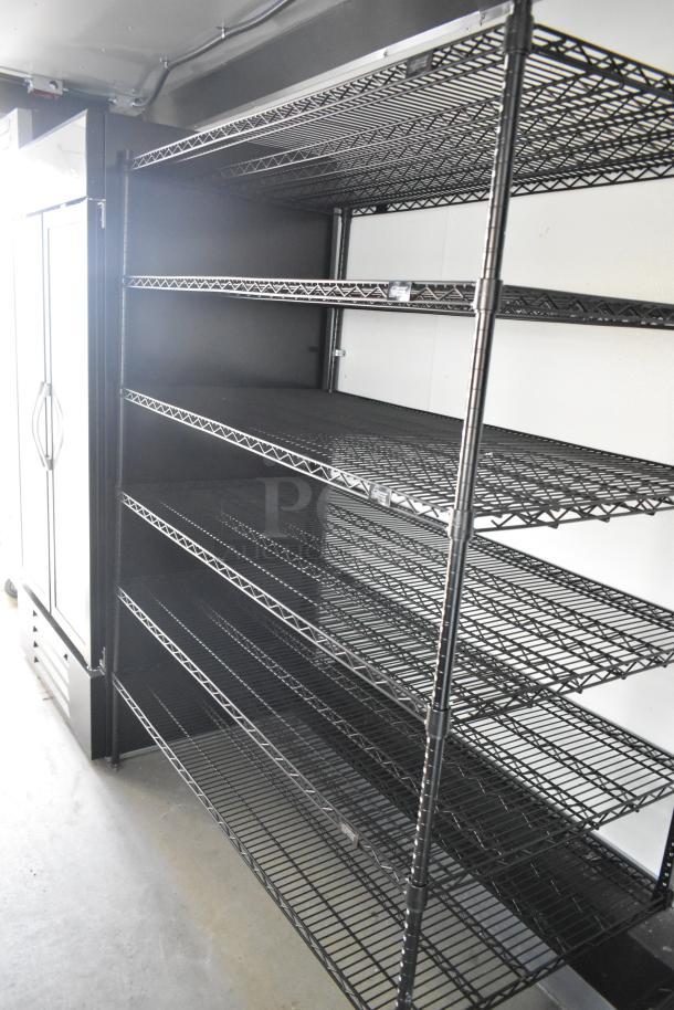 Alt Text: Black wire shelving in Nationwide 24' food trailer, next to a large white commercial cooler. New condition, fully equipped.