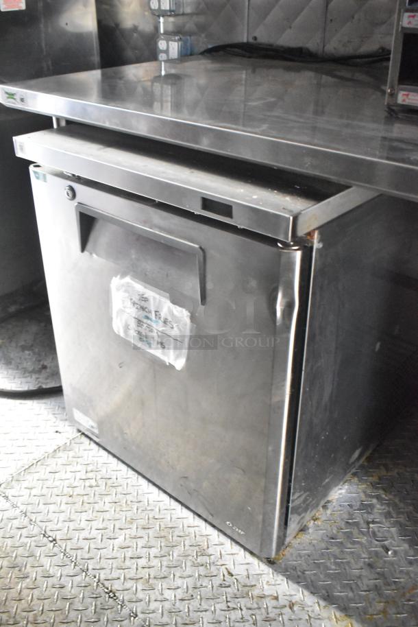 Commercial stainless steel under-counter refrigerator, dented door, interior shelf, taped label reads "Fridge/Freezer," power cord visible.