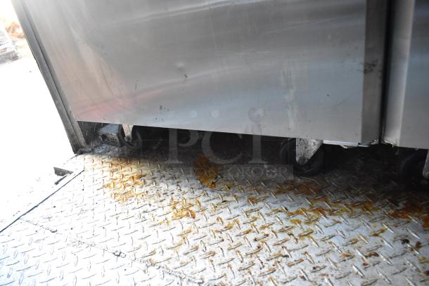 Stainless steel industrial refrigerator on casters, showing signs of wear. Diamond plate flooring beneath. No visible labels.