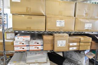 Sixteen unopened boxes containing Dual Lite LED emergency light back boxes, double remote heads, and exit signs on metal shelves. Tags show "LED Quantum" branding.