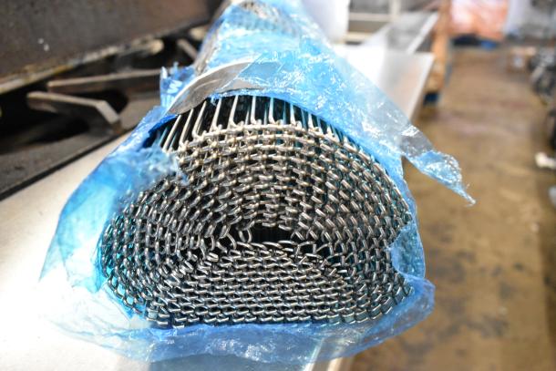 Brand new metal conveyor belt, rolled, showing slight scratches. Wrapped in blue plastic, revealing durable mesh construction.