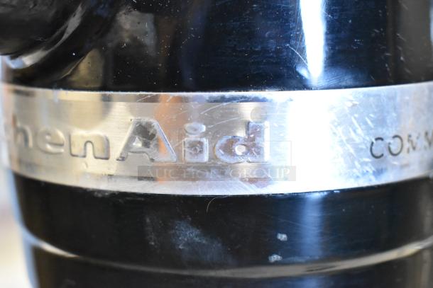 Close-up of a scratched KitchenAid KSM8990OB mixer label, showing "KitchenAid" text on a metal band. Black finish visible.