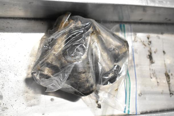 A metal component wrapped in a clear plastic bag, with greasy residue, on a metal surface.