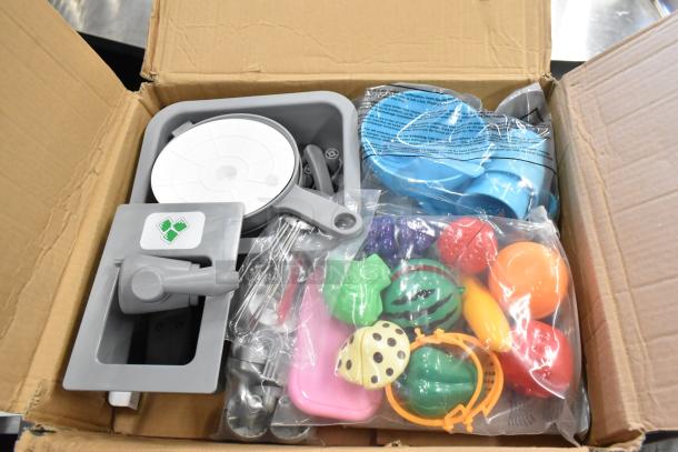 Box of cuttable fake toy food and accessories, including colorful fruits and kitchen tools, in new condition.