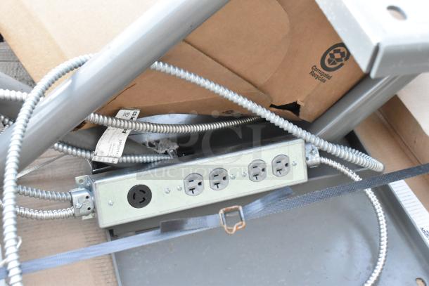 Electrical outlet strip mounted on a metal frame with attached cords, cardboard box in background.