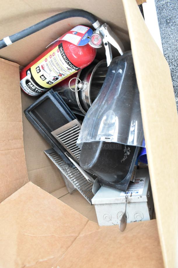 Cardboard box containing assorted items including a fire extinguisher, metal grates, and black plastic containers.