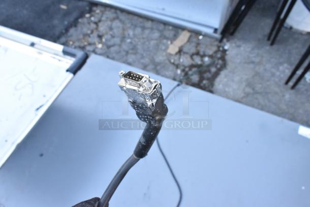 Gray panel with visible connector cable, close-up of a VGA connector, worn condition, on outdoor pavement background.