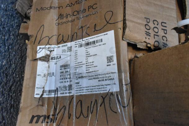 Cardboard box with visible damages and markings, containing a Modern AM243TP All-in-One PC. Label shows specs: Intel Core i7, 16GB RAM, 1TB HDD, Windows 11. Box labeled "MSI/LA," made in China.