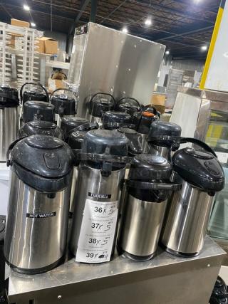 Commercial stainless steel insulated beverage dispensers, multiple units. Black lids, "Hot Water" label on some. Ideal for catering.