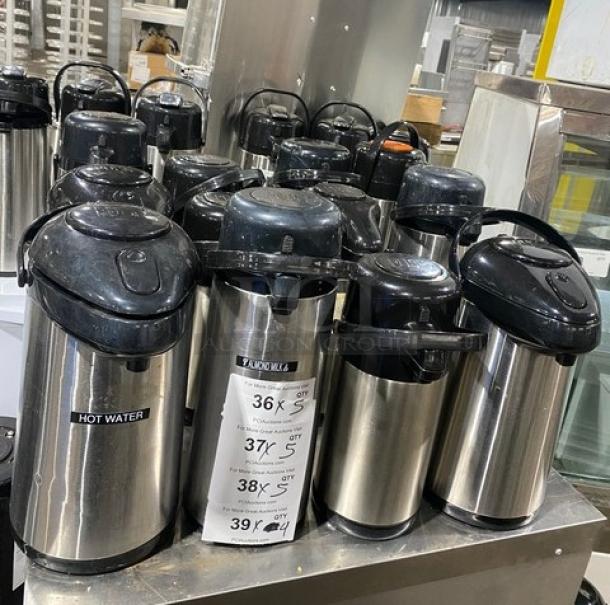Stainless steel insulated beverage dispensers on a metal counter. Multiple units labeled for hot liquids. Auction item.