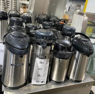 Stainless steel insulated beverage dispensers on a metal counter. Multiple units labeled for hot liquids. Auction item.