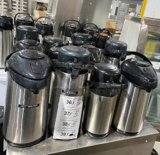 Commercial stainless steel insulated beverage dispensers, multiple units. Black lids, "Hot Water" label on some. Ideal for catering.