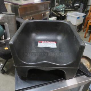 Black plastic booster seat with raised back and sides, featuring a visible warning label on the seating area. Used condition.