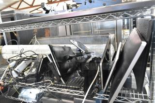 Eight assorted monitors including Toast POS, Elo POS, and Samsung S22D300HY, stacked on a metal shelf. Various cables visible.