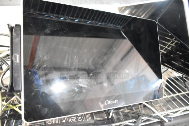 Eight electronic devices including Toast POS monitors, an Elo POS monitor, and a Samsung S22D300HY monitor on wire shelving.