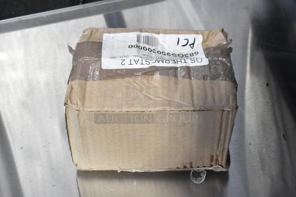 Brown box with shipping label, slightly dented. Label shows "GS1THERMOSTAT 2," indicating thermostat inside.