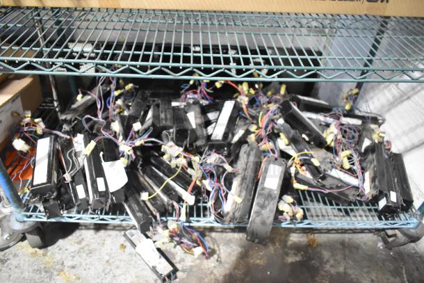 Lot of various electrical ballasts on a metal shelf. Items have wires and connectors, appear used, quantity unspecified.