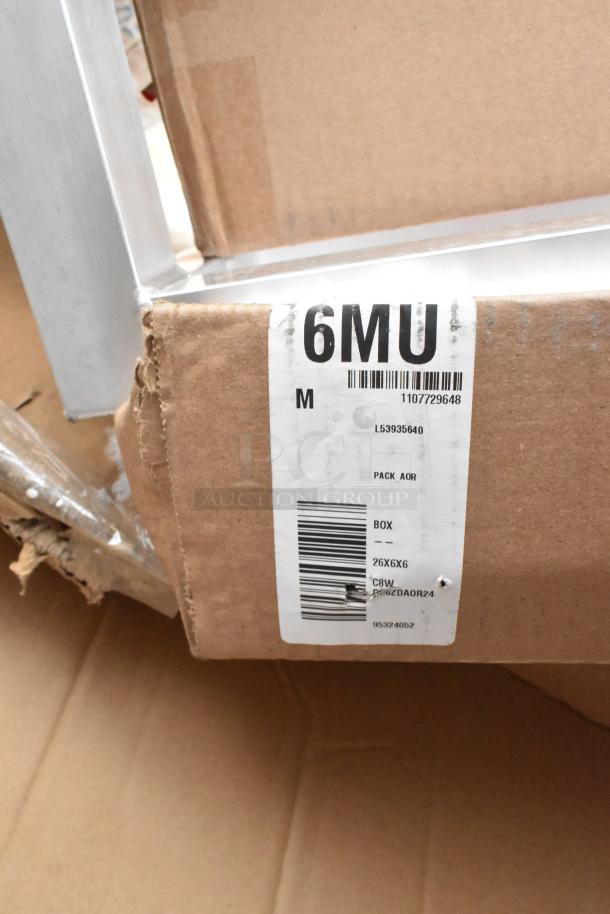 Cardboard box labeled "6MU M" with barcodes; contains new scratch and dent items including Lancaster Table & Seating folding tray stands.
