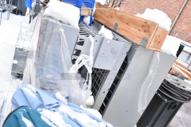 Gray metal pieces in a snowy outdoor setting, partially covered in plastic wrap, adjacent to wooden crate.
