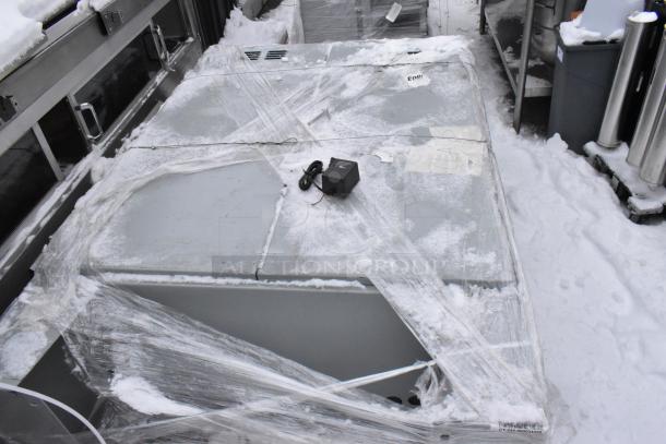 Large metal unit wrapped in plastic, covered with snow, showing a black electronic device on top. Marking indicates "Enge."