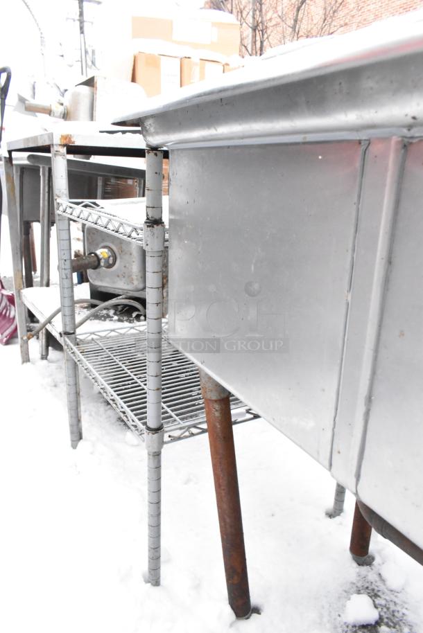 Stainless steel 2-bay sink with faucet, spray nozzle, and handles; minor rust on legs, outdoor setting.
