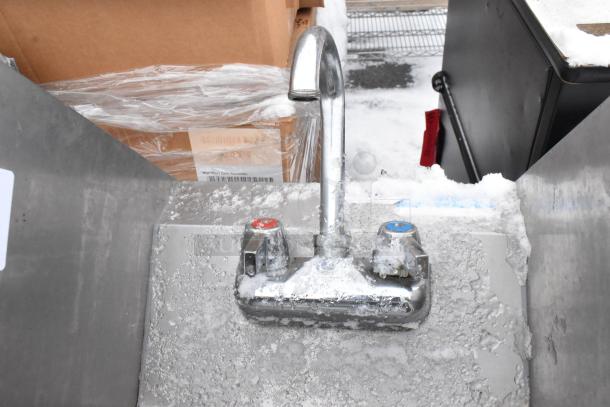 Stainless steel wall mount sink with faucet, handles, and side splash guards, shown with ice and snow.
