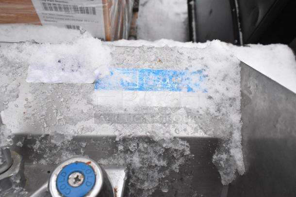 Stainless steel wall mount sink with faucet, handles, and side splash guards. Partial ID tag visible under snow.