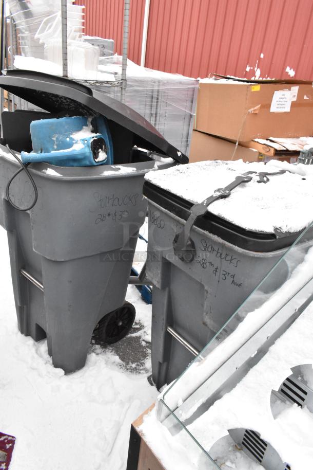 Two gray poly trash cans with lids and wheeled bases, containing a blue fan. Markings read "Starbucks 28th & 3rd Ave." Snow-covered.