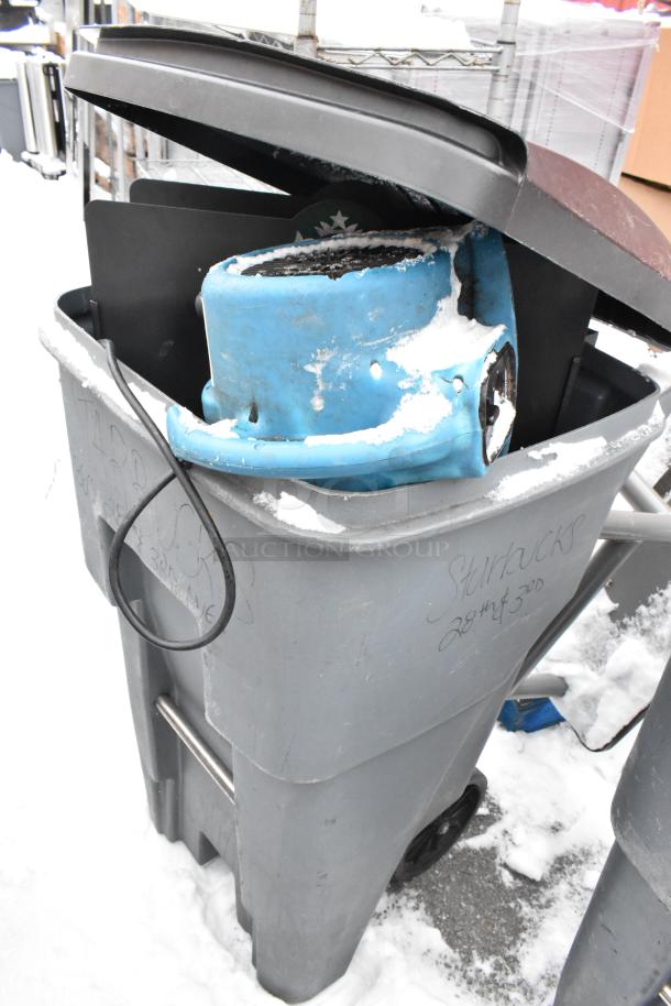 Gray poly trash can with lid, containing a blue fan and other items. Exterior markings visible. Snow on lid and rim.
