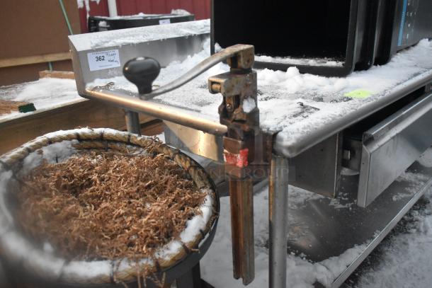 Stainless steel table with under shelf, includes mounted poly trash cans, metal clamp, visible label tag, snow-covered surface.