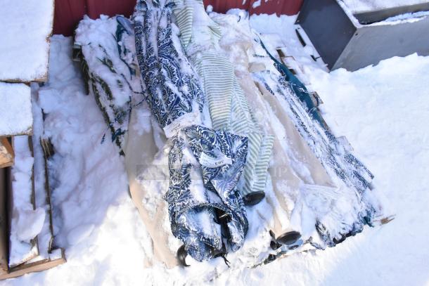 Lot of assorted patio umbrellas on a snowy surface, various patterns and colors, some tags visible, bundled together.