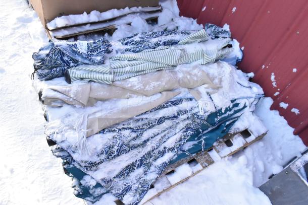 Lot of assorted patio umbrellas with snow-covered fabric, varied patterns and colors, stored on wooden pallets.