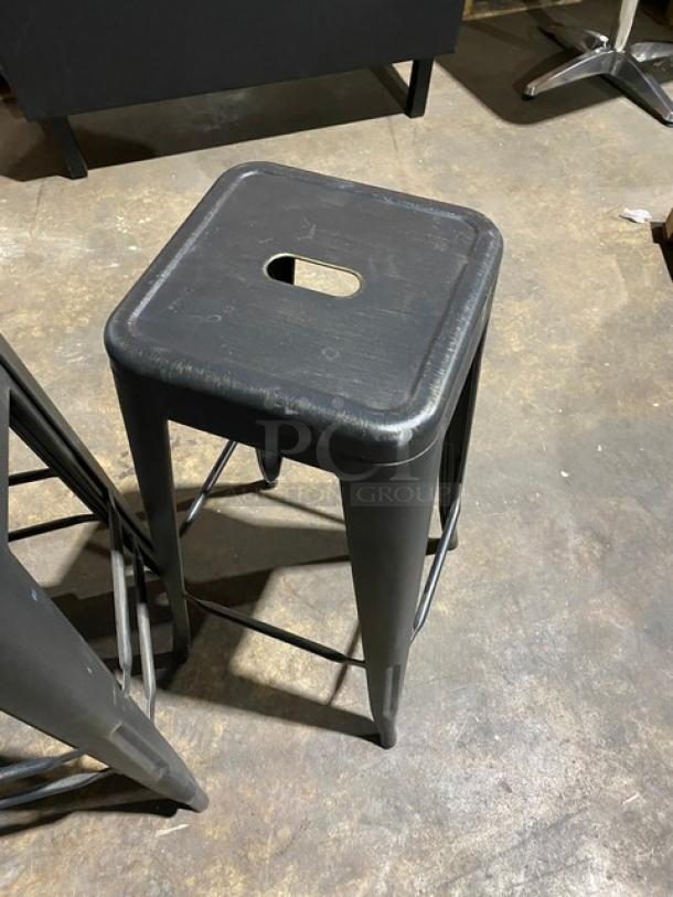 Brand new commercial black metal backless industrial-style stackable bar stool, featuring a square seat with cutout handle.
