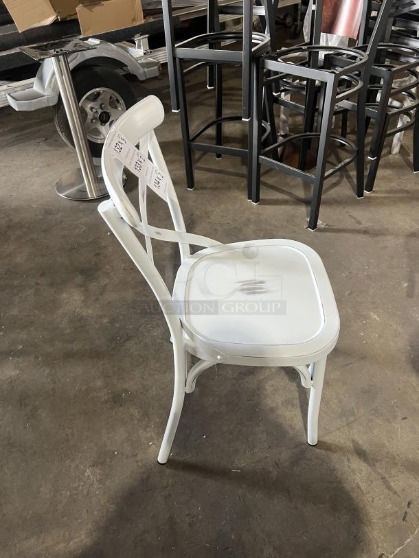 Commercial white cross-back dining chair, stackable design. Appears in good condition with minor scuffs. Auction tags visible.