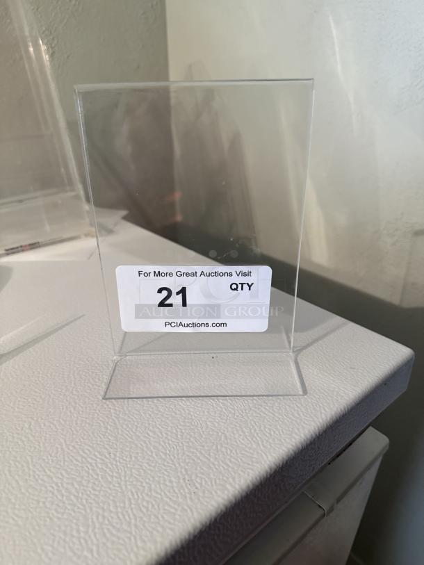 Six 5"x7" clear acrylic L-shaped sign holders. Perfect condition for menus or displays. Tagged with "21 QTY" from PCI Auctions.