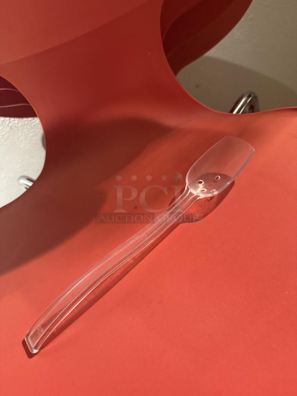 Clear slotted plastic serving spoon on red background, ideal for catering; slotted bowl design for easy draining.