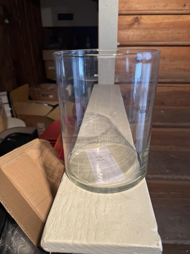 Large clear glass cylinder vase on shelf; versatile for decor, floral arrangements, or storage. Simple, elegant design.