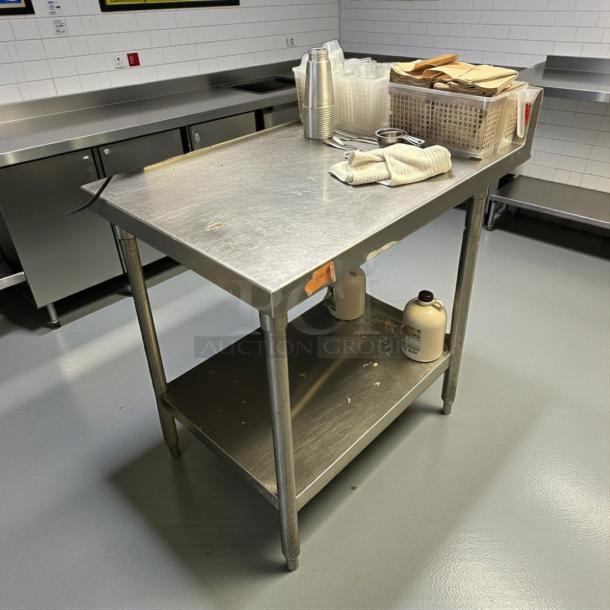 Omniteam stainless steel worktable, 24"x35", with backsplash. Features lower shelf, sturdy condition, minor wear visible.