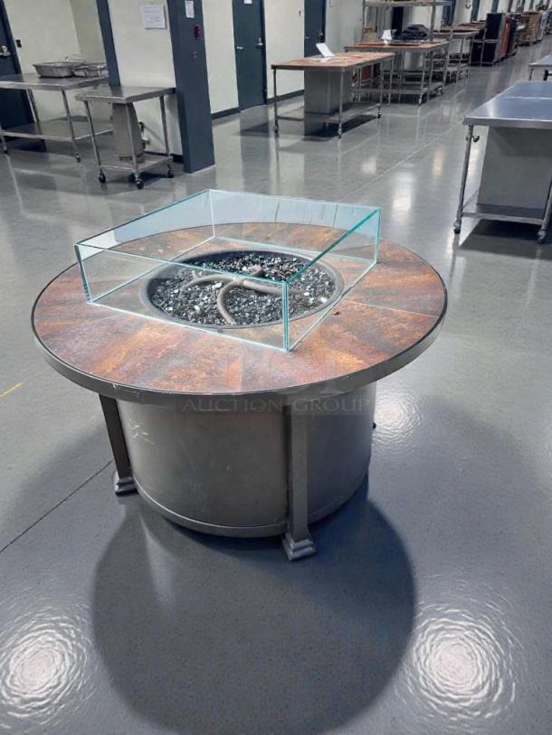 Round gas fire pit table in pristine condition by Outdoor Living. Features glass guard, natural gas/propane compatibility, inspected.