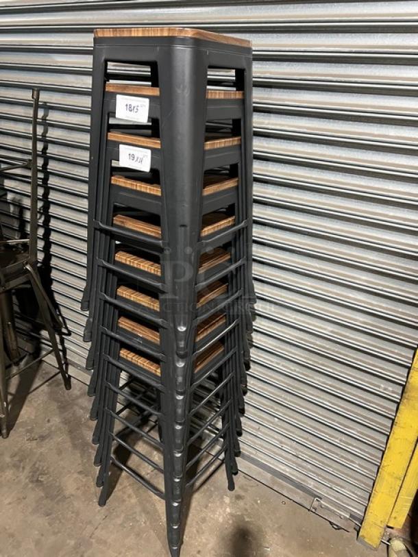 Stack of commercial black bar-height stools with wooden seats, stackable design, and tags indicating measurements.