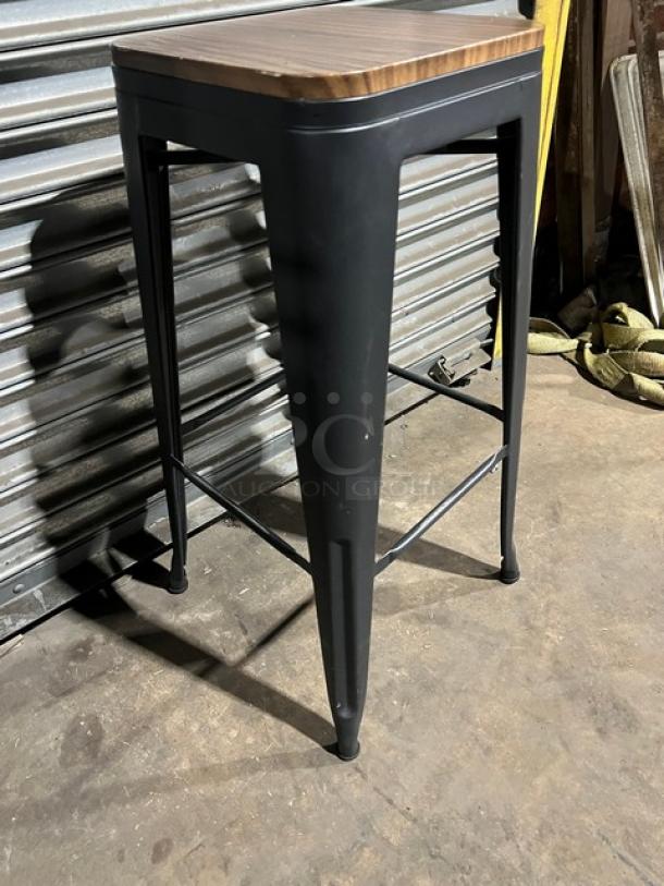 Commercial black stackable bar-height stool with wooden seat, metal frame, excellent condition, durable for heavy use.