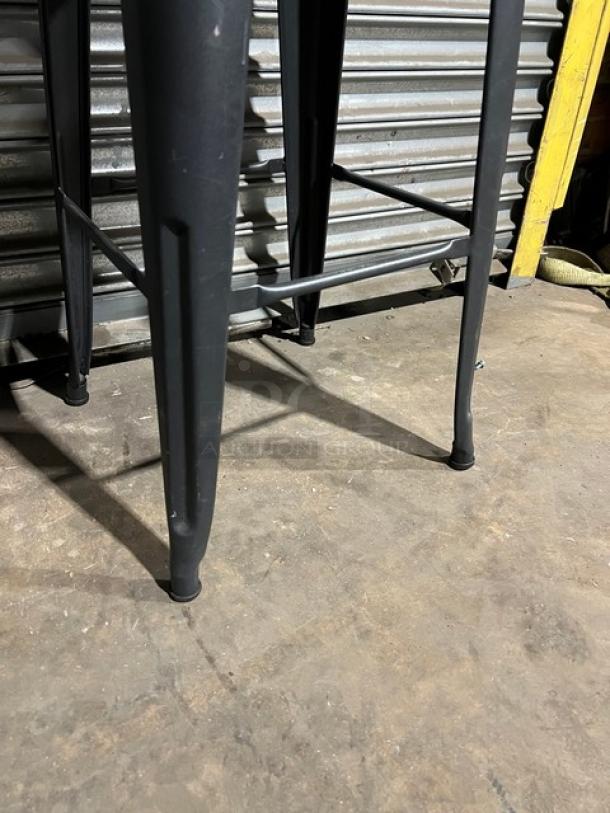 Commercial black stackable bar-height stool with metal legs, shown in good condition on a warehouse floor.