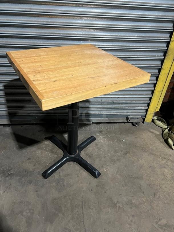 Square commercial dining-height table with a wood top and sturdy black metal pedestal base. Suitable for restaurants.