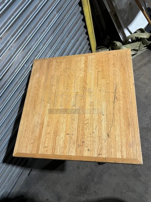 Square wooden commercial dining-height table with visible wear and scuff marks, showcasing a natural wood grain pattern.