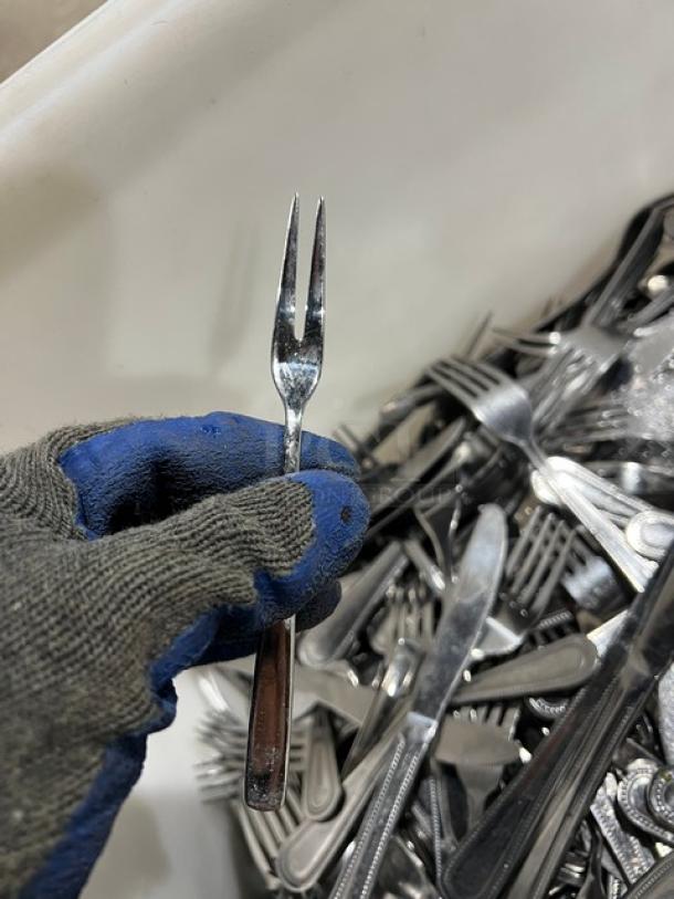 Stainless steel dining utensils assortment, including forks, knives, spoons in a bin. Gloved hand holding a small serving fork.