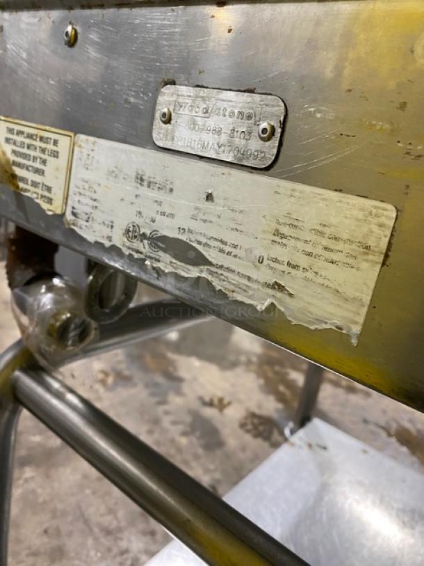 WoodStone commercial stainless steel natural gas countertop griddle with back and side splash, serial GP1B16MAY1704992. Features include single undershelf cart on casters. Condition shows visible wear with intact manufacturer labels.