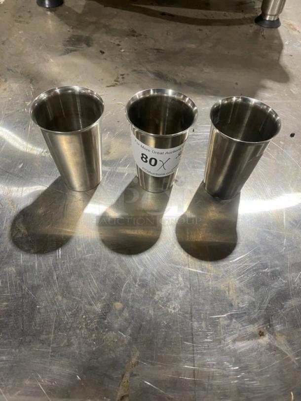 Three stainless steel malt shaker cups on a metal surface. One cup has an auction tag labeled "80X."
