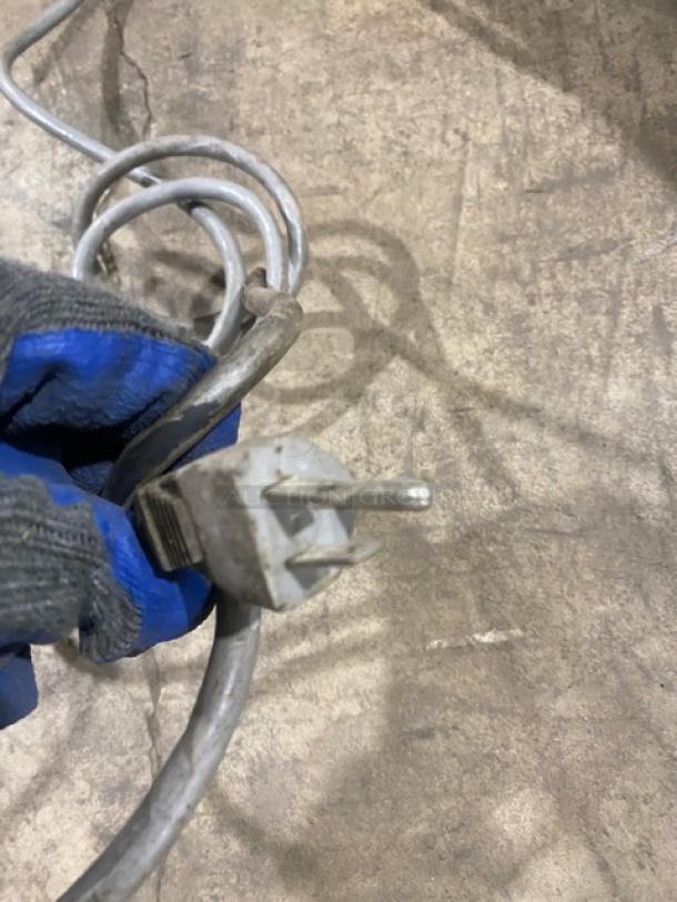 Close-up of an electrical plug on a coiled grey cord. Plug is slightly dirty. Surface in background is worn.