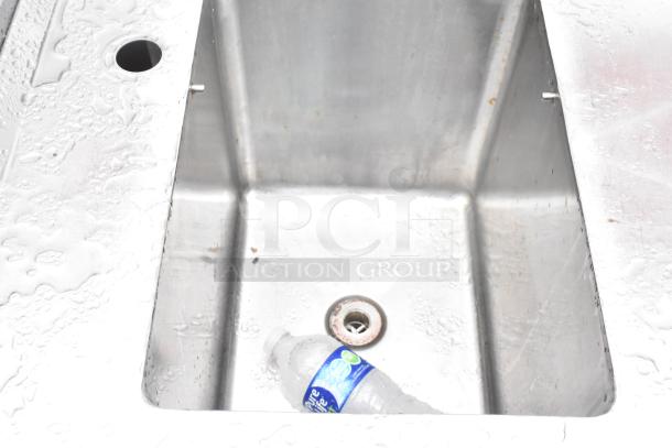 Stainless steel commercial single bay sink with backsplash and under shelf, showing minimal wear, drain visible.